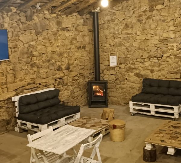 Interior rústico de La Palleira con paredes de piedra, estufa de leña, sofás y mesas de madera, espacio acogedor en Solpor Camperpark.