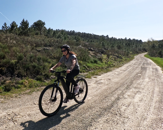 Mujer pedaleando en una e-bike por un camino de tierra entre pinos en la Ribeira Sacra durante una ruta guiada.
