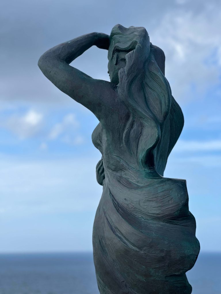 Detalle de la escultura A Espera de Laxe con el cielo y el mar al fondo