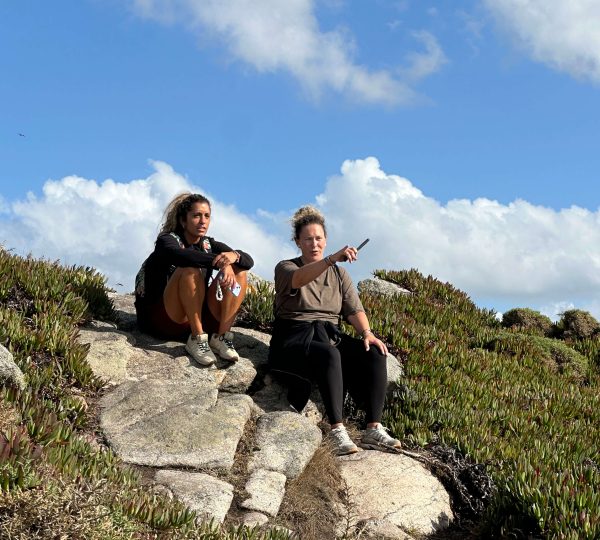 Dos mujeres descansan sobre unas rocas durante una ruta por la costa